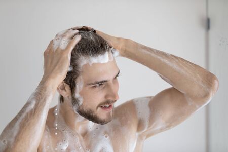 Close up of young naked caucasian man wash clean hair with foamy natural shampoo in bathroom,...