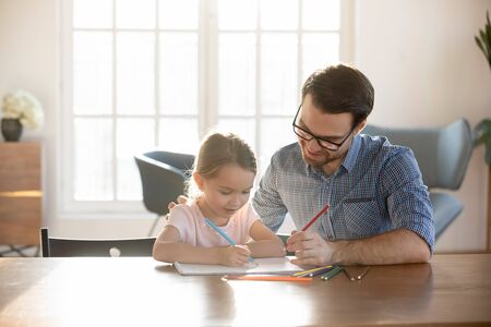 Loving young father sit at desk studying drawing with small preschooler daughter, caring happy dad have fun painting picture with little girl child, enjoy leisure family weekend at home togetherの写真素材