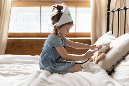 Cute little girl dressed as doctor playing with toy, using stethoscope, checking listening to heart beat, pretty preschool child pretending pediatrician, sitting on cozy bed in bedroomの写真素材