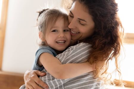 Head shot portrait close up loving mother and adorable little daughter cuddling, smiling cute preschool girl looking at camera, hugging caring young mum, family enjoying tender momentの写真素材