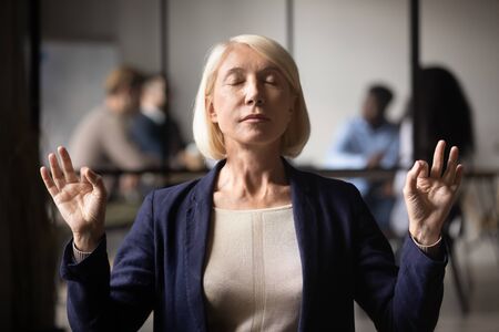 Calm middle aged businesswoman with closed eyes meditating at work, mature female employee practicing breathing exercise in office, stress relief concept, keep clam, hands in mudra gestureの写真素材