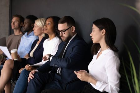 Bored diverse people candidates waiting for job interview, sitting in row in queue in office room, using gadgets, holding resume documents, human resources, recruitment and employment conceptの写真素材