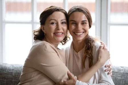Portrait of smiling mature Caucasian mom and grownup daughter hug relaxing at home together, happy...