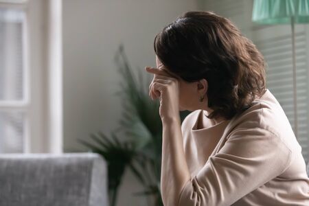 Upset worried mature woman sit on couch feel lonely abandoned missing or mourning at home, unhappy anxious middle-aged female look in distance thinking or pondering, elderly solitude conceptの写真素材