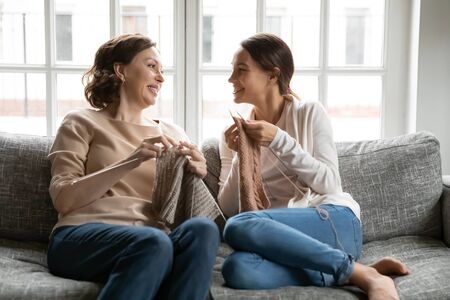 Smiling middle-aged mother and adult daughter relax on sofa at home knit with needles together, happy mature mom and grownup millennial girl child engaged in hobby activity, yarn on weekendの写真素材