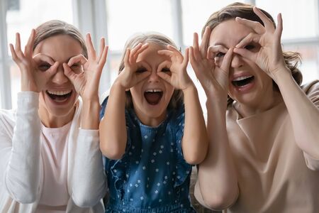 Portrait of overjoyed three generations of women have fun make funny faces gesture relaxing at home together, happy little girl with young mom and mature grandmother enjoy family weekend reunionの写真素材