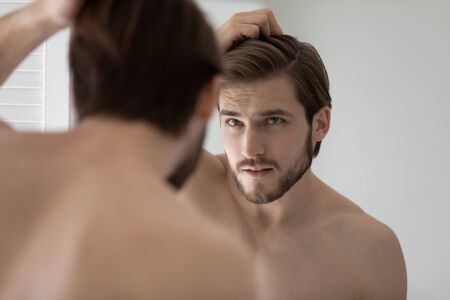 Anxious young Caucasian man look in mirror in bathroom touch check head worried about dandruff, concerned millennial male frustrated about hair loss, receding hairline, haircare, beauty conceptの写真素材
