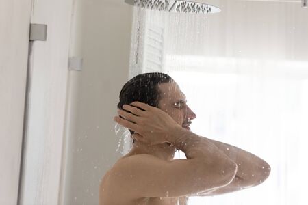 Naked millennial man stand under water flow washing in morning in home bathroom, young caucasian...