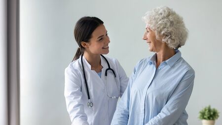 Caring young female caregiver in white coat smiling looking at old woman patient showing her support friendly attitude warm relation, nursing and healthcare, social homecare of elderly people conceptの写真素材