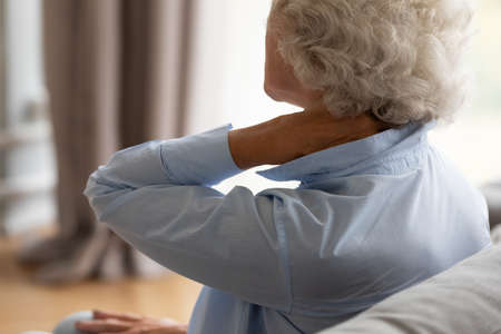 Tired elderly 70s woman sit on sofa massaging stiff sore neck tensed muscles reduces painful feelings, shoulder back pain fibromyalgia osteoarthritis senile chronic diseases concept close up rear viewの写真素材