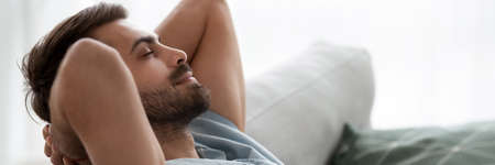 Calm millennial man leaned on couch put hands behind head closed eyes resting in living room, enjoy lazy day, breath fresh air feels good, close up photo. Horizontal banner for website header designの写真素材