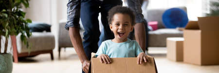 African dad play with excited toddler son riding seated inside of box in moving day at new home, little kid enjoy game with daddy, relocation concept. Horizontal photo banner for website header designの写真素材