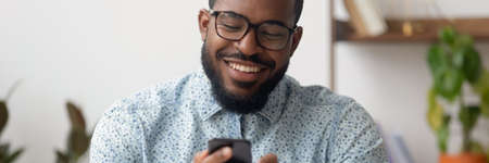 Smiling African businessman sit at workplace holding mobile phone, texting sms, browsing internet. Distant communication, virtual chat usage concept. Horizontal photo banner for website header designの写真素材