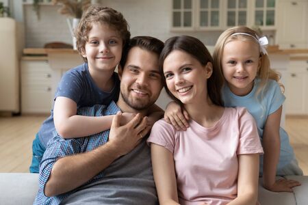 Portrait of happy young Caucasian family sit on couch in kitchen posing with little preschooler kids, smiling mother and father hug embrace small daughter and son relaxing together on weekend at homeの写真素材