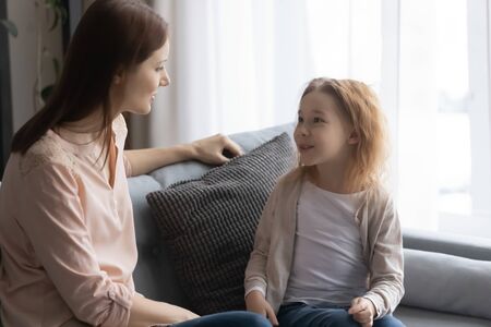 Loving young Caucasian mother sit rest on sofa talking chatting with small preschooler daughter, little girl child relax on couch at home, speak enjoy weekend with mom or nanny, sharing secretsの写真素材