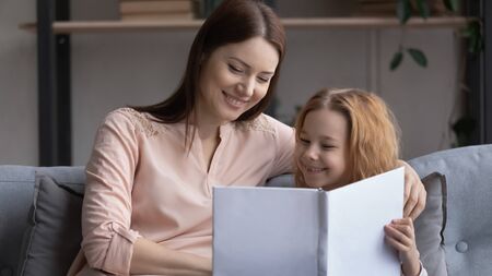 Smiling young Caucasian mom and little preschooler daughter sit on sofa at home reading book...