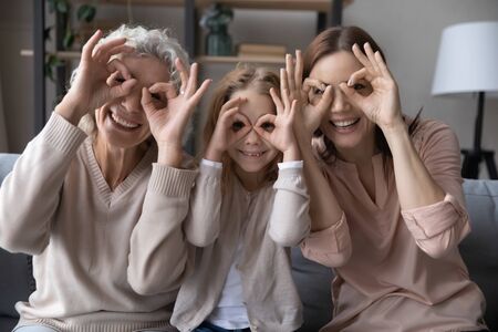 Portrait of overjoyed three generations of women have fun make funny facial gestures relax at home together, smiling small girl with young mom and mature grandmother play enjoy family weekend reunionの写真素材