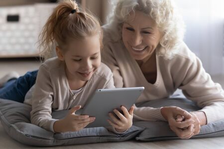 Playful happy elderly grandmother and little granddaughter relax lying on floor at home watching video pad gadget, smiling mature grandparent have fun play using modern tablet with small grandchildの写真素材