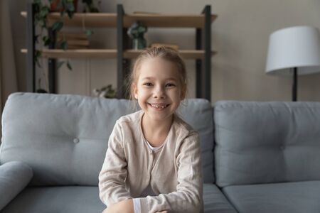 Headshot portrait of smiling little preschooler girl sit at couch in living room talk on video call, happy small child have webcam online conference or conversation at home, virtual technology conceptの写真素材