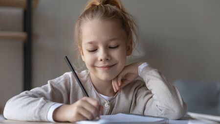 Close up of smiling small girl sit at desk handwriting in notebook, doing homework, happy motivated...
