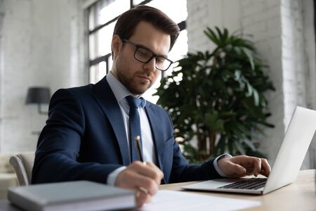 Concentrated Caucasian young businessman sit at desk in office work on laptop making notes, serious...
