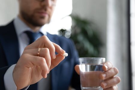 Close up of male worker hold glass of water and pill think of taking antibiotic or aspirin, unhealthy man employee feel unwell have medicines daily dose of vitamins at workplace, healthcare conceptの写真素材