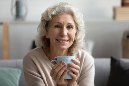 Close up portrait of happy senior 50s woman relax on couch at home drink hot tea or coffee from cup, smiling elderly female look at camera posing, rest on sofa in living room enjoying weekend indoorsの写真素材