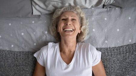 Top view of excited mature woman lying in bed at home have fun laughing and joking, overjoyed happy elderly grandmother feel optimistic about good maturity, relax rest in comfortable bedroomの写真素材