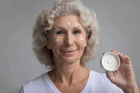 Headshot portrait of elderly 60s woman apply moisturizing anti-age facial cream, recommend good beauty product, happy mature 50s grandmother use nourishing anti-wrinkle face mask, skincare conceptの写真素材