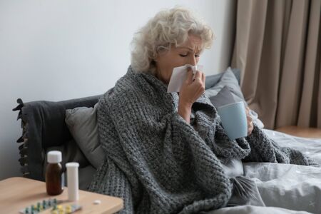 Sick older woman sit in bed blow running nose, suffer from flu or cold at home, ill mature female feel unhealthy struggle with influenza, sneeze or snuffle under blanket, elderly healthcare conceptの写真素材