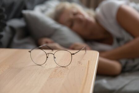 Close up of round glasses lying on bedside table in bedroom, senior woman sleep in bed in background, mature grandmother take off spectacles taking nap, elderly eyesight problem correction conceptの写真素材