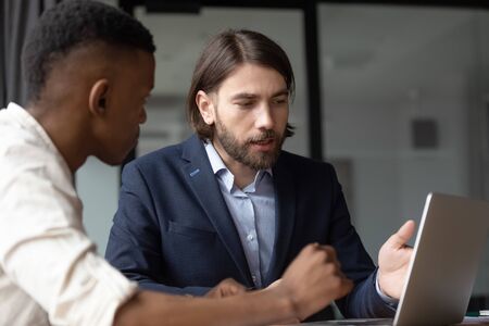 Mentor teach intern explains corporate app sitting at desk in office. Caucasian and African businessmen negotiating, client and manager communicating, make offer, selling services to customer conceptの写真素材