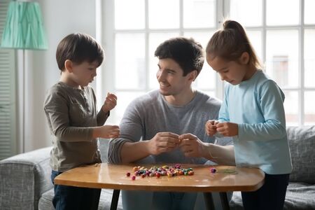 Loving young Caucasian father string colorful wooden beads on thread making bracelets accessories with preschooler kids, happy dad have fun engaged in creative artistic activity with small childrenの写真素材