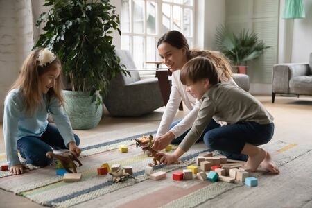Loving young mom sit on floor in living room have fun playing with excited little children with toys, playful caring Caucasian mother or nanny engaged in funny game with small kids in living roomの写真素材