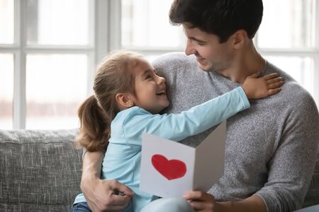 Cute little girl hug cuddle happy young father present handmade postcard for birthday or anniversary, smiling small daughter embrace dad greeting congratulating at home, show love and gratitudeの写真素材
