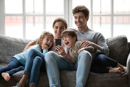 Overjoyed young family with small children sit relax on sofa at home laugh watching funny video on smartphone together, happy Caucasian parents rest with little kids using modern cellphone gadgetの写真素材