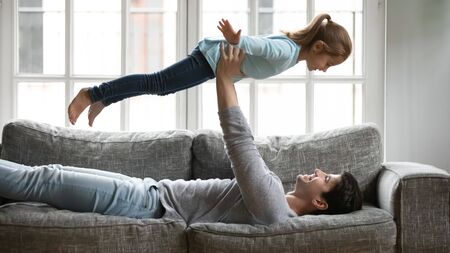 Smiling young father lying on couch in living room hold in arms imitate plane with little preschooler daughter, happy Caucasian dad have fun playing on sofa with overjoyed small girl child at homeの写真素材