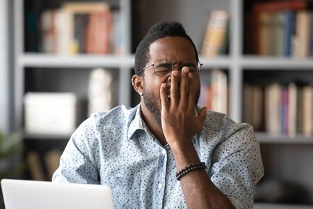 African 30s man sitting at desk in front of laptop, yawns covers mouth with palm feels unmotivated at workplace. Concept of drowsiness, overworked freelancer, boring job, early morning Monday conceptの写真素材