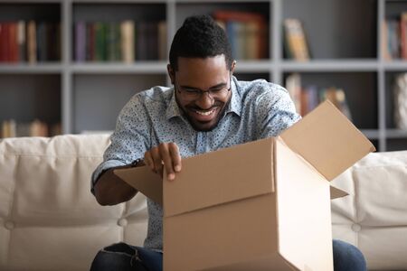 Interested African guy in eyeglasses sit on couch at home unbox long-awaited parcel. Happy internet shopper got quality product from online store, delivery service trusted transporting company conceptの写真素材