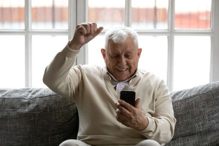 Overjoyed smiling mature man reading good news in smartphone, looking at phone screen, showing yes gesture, sitting on couch, excited happy elderly male celebrating success, online lottery winの写真素材