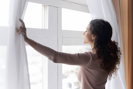 Close up smiling woman opening curtains in morning, enjoying sunlight, standing at home, happy overjoyed young female looking out window, dreaming, starting new day, planning good futureの写真素材