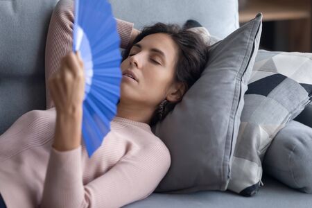Exhausted overheated woman waving blue paper fan close up, lying on couch, relaxing on soft pillows, enjoying fresh air, tired young female suffering from heating, hot summer weather or feverの写真素材