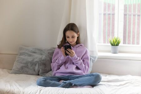 Smiling teenager relax on comfy bed at home playing or browsing internet on cellphone, happy teenage girl sit rest in bedroom texting messaging online on smartphone, communication, technology conceptの写真素材