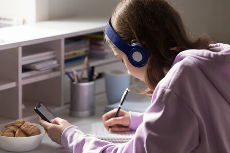 Concentrated teenage girl in headphones sit at desk at home write in notebook prepare homework using...