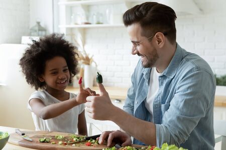 Caucasian daddy African daughter having fun put on fingers cucumber and paprika pieces like puppet toys making up funny vegetable stories while preparing vegetarian natural salad together in kitchenの写真素材