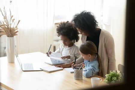 African mom of two multiethnic school age daughters do lesson homework helps them. American and Caucasian girls siblings writing on workbooks study with teacher at home. Homeschooling tutoring conceptの写真素材