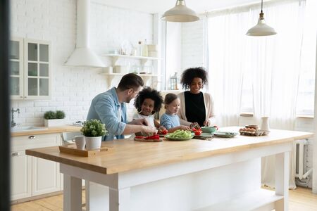 Happy mixed race multinational family with daughters prepare vegetable organic fresh salad, enjoy pastime cooking process in cozy kitchen, healthy eating, parents teach kids, weekend at home activityの写真素材