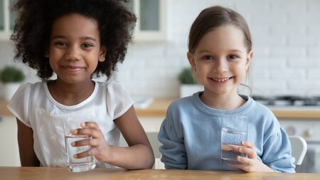 Close up horizontal image two school age girls african caucasian little adorable kids sit in kitchen holding glasses with still clear water look at camera. Healthy lifestyle, natural hydration conceptの写真素材