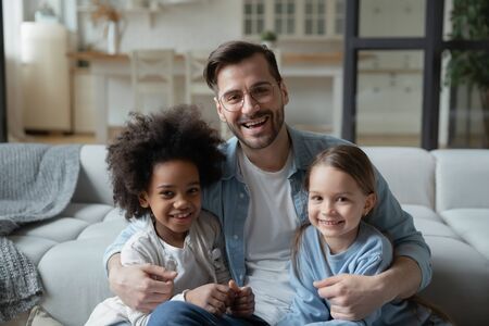 Father Day celebration, happy fatherhood multiracial family portrait concept. Caucasian stepdad sit on sofa hugs european mixed-race african daughters people smile pose for camera photo shoot at homeの写真素材