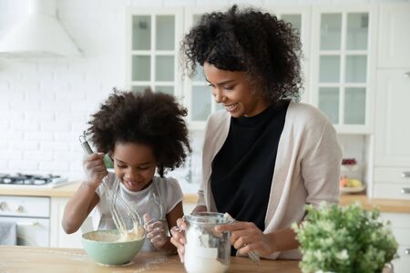Little daughter helper and African mother cooking together in kitchen, kid girl preparing cake or...
